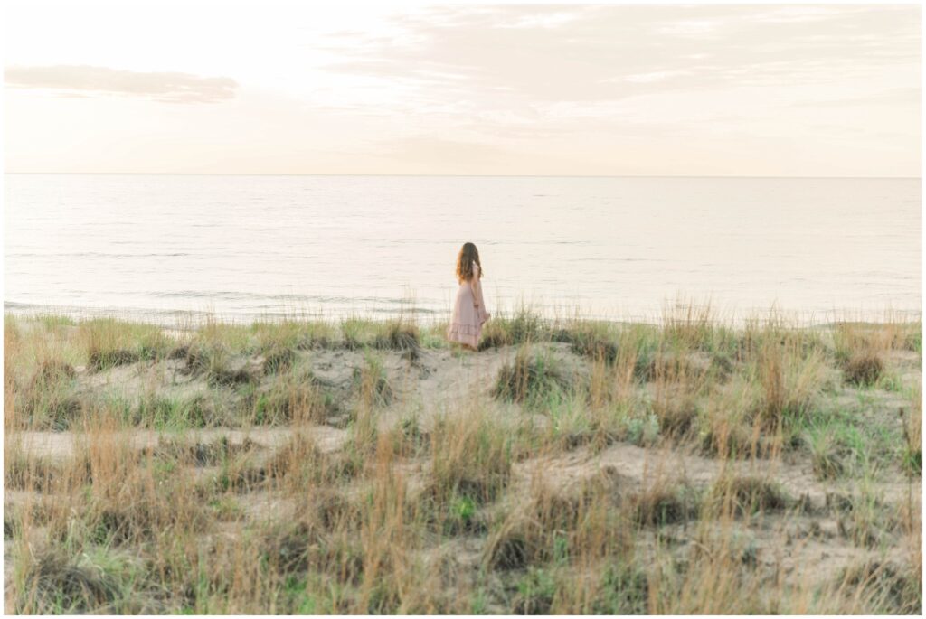 Indiana dunes family photos: young girl in a pink dress looks out over lake michigan surrounded by beach grass and sand. A soft golden sunset gives everything a glow.
