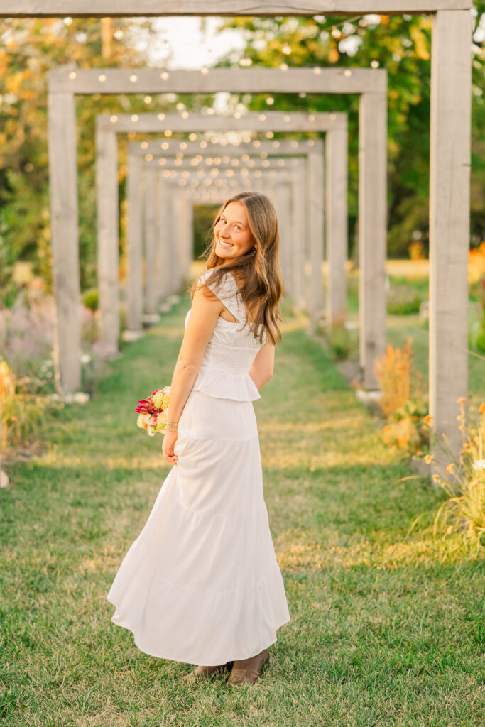 High School senior girl in white dress with a bouquet of flowers and twinkle lights behind. 
