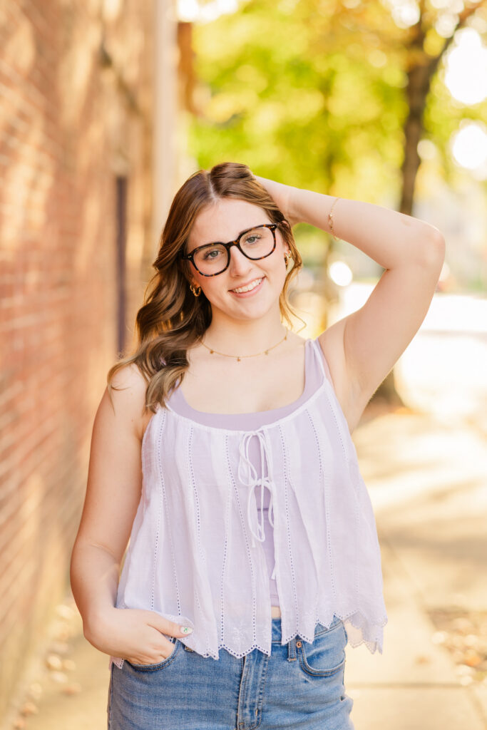 Senior girl with hand in her hair posing in a charming downtown photo location with brick walls and the glowy sun behind her.