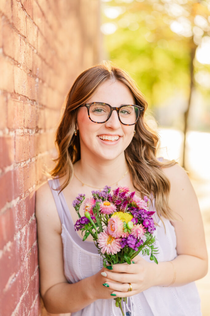 Senior girl with glasses holding a bouquet of colorful flowers and leaning against a wall.