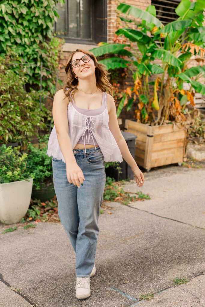 Senior girl wearing jeans and a casual top walking towards the camera with green plants in the background.