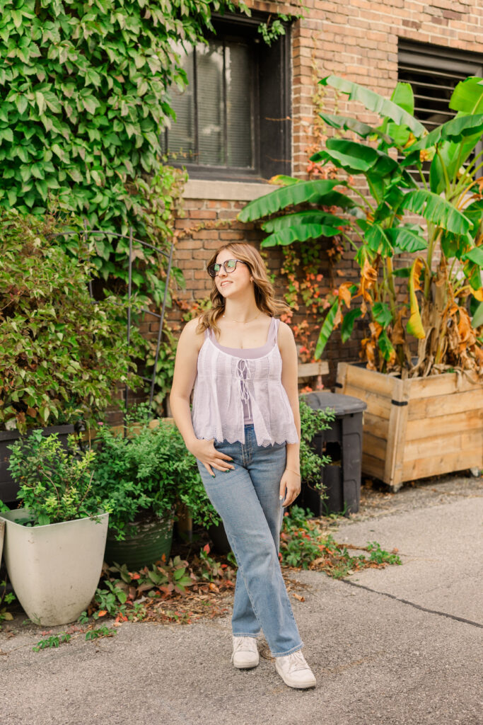 Senior girl with jeans and a casual top posing in an urban downtown alleyway with red brick walls and greenery in pots.